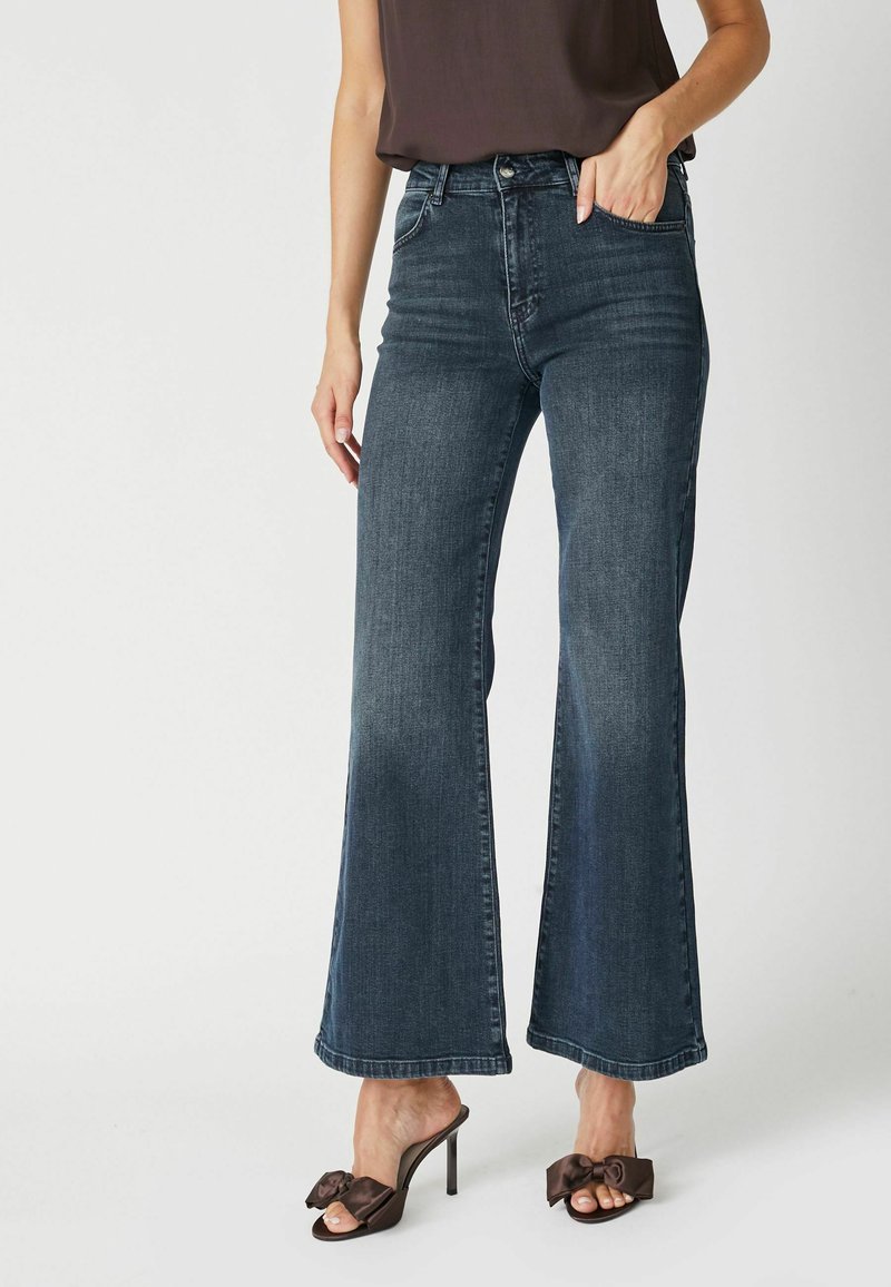 Jeans de mezclilla de tiro alto en un azul oscuro y de pierna ancha. Combinados con sandalias de tacón marrones con un detalle de lazo.