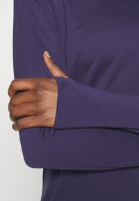 Pitkähihaista urheilu-toppi syvän purppuran värisessä kankaassa, jossa on tiukka rannekura. Materiaali näyttää sileältä ja siinä on minimaalinen tekstuuri.