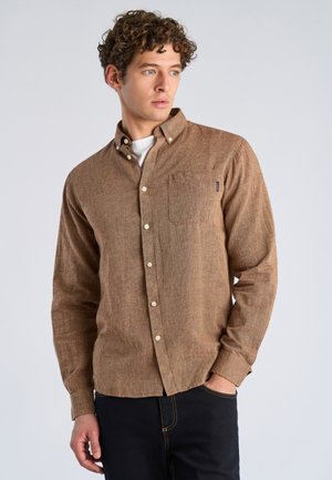 Junger Mann mit lockigem Haar, der ein braunes Hemd über einem weißen T-Shirt trägt, mit einer Hand in der Tasche stehend und seitlich blickend.