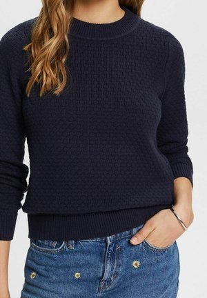 Granatowy sweter dziany z fakturowanym wzorem, okrągłym dekoltem i ściągaczami przy mankietach. W zestawie z jasnoniebieskimi szortami dżinsowymi ozdobionymi kwiatowymi akcentami.