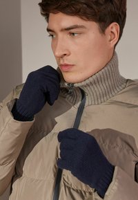 Granatowe dzianinowe rękawiczki z prążkowanymi mankietami noszone z beżową pikowaną kurtką mającą wysoki kołnierz i zamek błyskawiczny. Miękka tekstura i nowoczesny design.