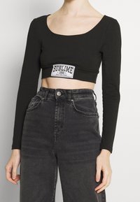 Black long-sleeve crop top featuring a white graphic label reading "SUBLIME 1995" on the front. Paired with high-waisted black denim jeans.