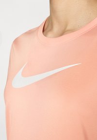 Vaaleanpunainen urheilupaita, joka on valmistettu sileästä kankaasta, ja etuosassa on valkoinen Nike-logon swoosh ja klassinen pyöreä pääntie.