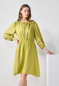 Hellgrünes Kleid aus strukturiertem Stoff, mit elastischem Bund und Puffärmeln mit gerafften Details. Der Ausschnitt ist mit einer Schleife verziert.
