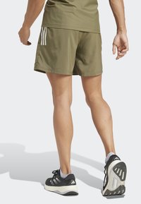Person wearing olive green sports shorts and shirt with black and white Adidas running shoes, shown from the back mid-stride.