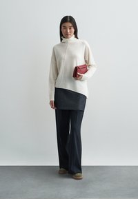 Cream-colored turtleneck sweater with a diagonal seam, paired with a dark denim skirt and loose black trousers. Holding a red clutch.