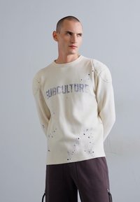 Felpa a costine color crema con stampa "SUBCULTURE", caratterizzata da macchie di vernice e dettagli sulle spalle, abbinata a pantaloni marroni.