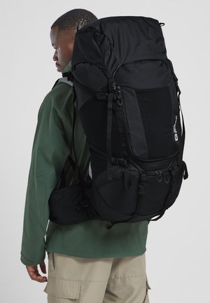Schwarzer Wanderrucksack mit strukturiertem Design, mehreren Riemen und Taschen, aus strapazierfähigem Material gefertigt. Getragen von einer Person in einer grünen Jacke.