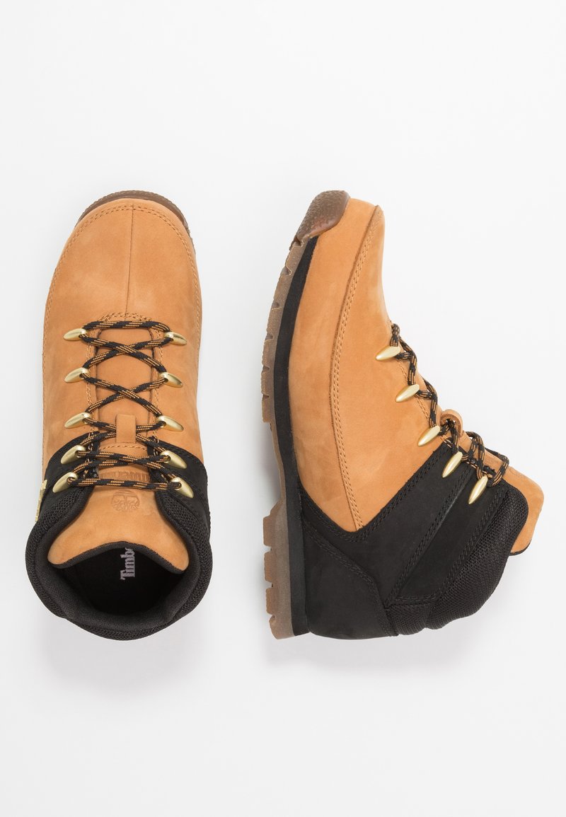 Botas de senderismo de ante en tono marrón con acentos negros, que presentan ojales dorados, cordones redondos y suelas de goma texturizadas para mayor tracción.
