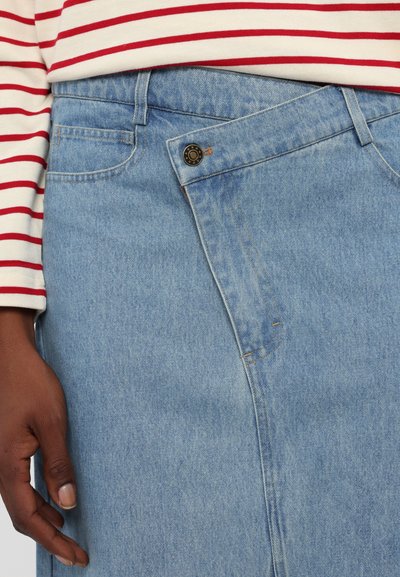 Svetlo modra jeans krilo z ukrivljenim sprednjim dizajnom, šivano detajli in eno kovinsko gumbom na pasu. Črtasta srajca delno vidna.