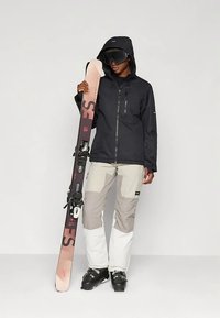 Cilvēks, kurš ģērbjas melnā slēpošanas jakā, gaišpelēkās un baltās biksēs, slēpošanas zābakos un brillēs, tur pa pāri rozā un melnā krāsā gadījumiem slēpes vertikāli.