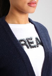 Dunkelblauer Cardigan mit weicher Textur, getragen über einem weißen T-Shirt mit einem pailettenverzierten "REAL"-Design in Schwarz und Silber.
