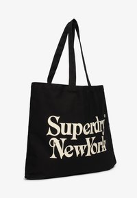 Czarna torba na ramię z długimi uchwytami, z dużym kremowym napisem "Superdry New York" z przodu. Prosty design, solidny materiał.