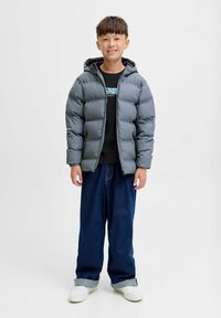 Dječak stoji, obučen u sivi puffer kaput, crnu majicu s printom, široke plave traperice s manžetama i bijele tenisice na jednostavnoj svijetloj pozadini.