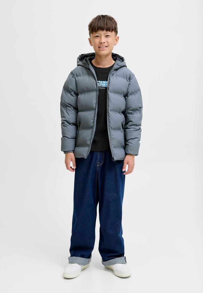 Dječak stoji, obučen u sivi puffer kaput, crnu majicu s printom, široke plave traperice s manžetama i bijele tenisice na jednostavnoj svijetloj pozadini.