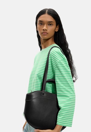 Kobieta z długimi czarnymi włosami, ubrana w zieloną koszulę w paski, niosąca na ramieniu dużą czarną skórzaną torbę typu tote na jednolitym tle.