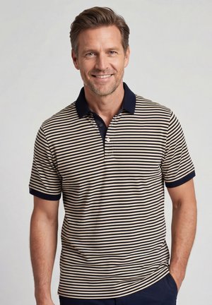 Lachende man met lichtbruin haar draagend een kortemouw navyblauwe en beige gestreepte polo, staand met één hand in de zak.