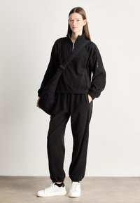 Femeie în picioare, îmbrăcată cu un pulover negru cu fermoar, pantaloni negri asortați, adidași albi și purtând o geantă crossbody neagră pe umărul drept.