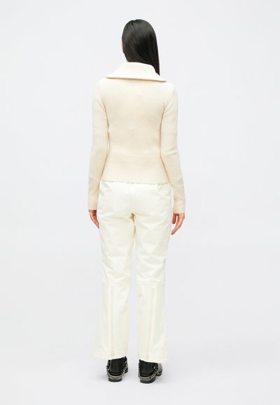 Strikket kremhvit genser med vid krage, tettsittende kropp og ribbet kant, kombinert med hvite vide bukser med glidelåser på ermene og svarte støvler.