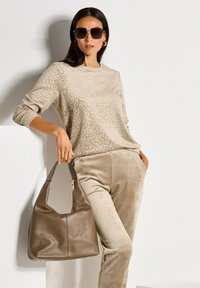 Beiger Pullover mit dezentem Leopardenmuster, kombiniert mit beigen samtigen Hosen. Eine braune Leder-Hobo-Tasche mit strukturiertem Finish wird in der Hand gehalten.