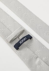 Ceinture en tissu gris à motifs présentant une texture tissée, avec une étiquette marine portant le mot "JOOP!" fixée. Des motifs floraux subtils rehaussent le design.