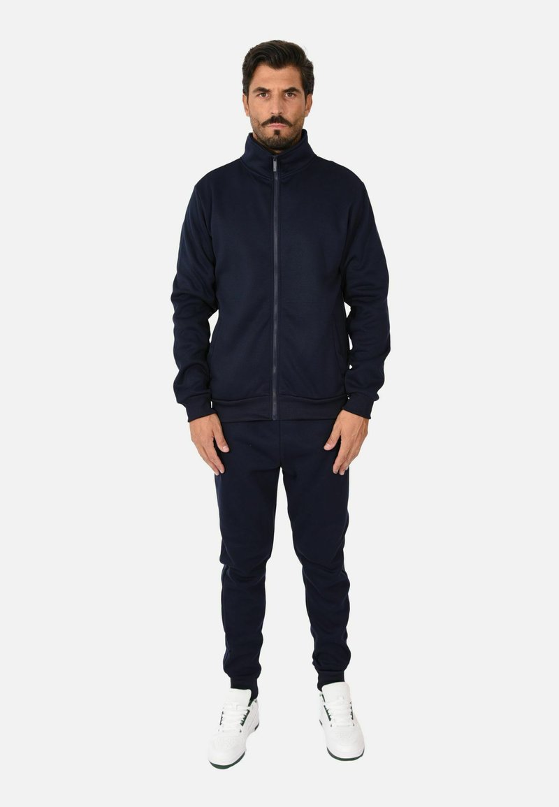 Tuta navy realizzato in tessuto morbido, composto da una giacca con zip e colletto alto e pantaloni affusolati coordinati, abbinati a sneaker bianche.
