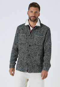 Sako vzoru rybej kosti v čierno-bielej farbe, so zapínaním na gombík, veľkým golierom a dlhými rukávmi, nosené cez červenú košeľu.