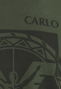 Tmavě zelené bavlněné tričko s velkým černým grafickým potiskem a textem "CARLO" v tučném písmu blízko ramene.