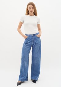 Jeans in denim blu a gamba larga con vita alta, dotati di chiusura con bottone e tasche anteriori, abbinati a una T-shirt bianca aderente a coste.