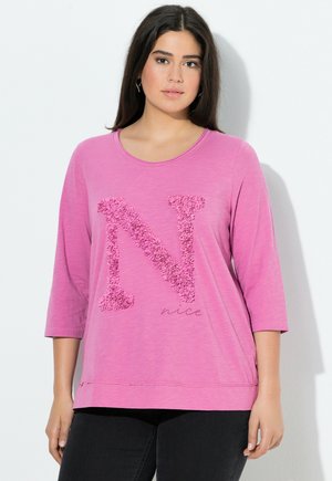 Chemise rose à manches longues ornée d'une lettre "N" texturée et du mot "nice" en dessous. Design décontracté avec un col rond et une coupe ample.