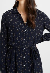 Granatowa sukienka z długim rękawem, w kwiatowy wzór, z guzikami z przodu, kołnierzykiem i paskiem w talii. Wykonana z lekkiego materiału, ma delikatny blask i fakturę.