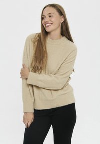 Beige, nadýchaný svetr s kulatým výstřihem, dlouhými rukávy a texturovaným povrchem. Kombinován s černými kalhotami. Jednoduchý design a měkký materiál.
