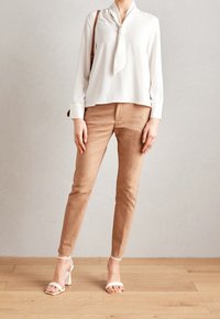 Žena v bílé dlouhé blůze s uvázaným límcem, béžových úzkých kalhotách, na bílé vysoké sandály a nese hnědou kabelku přes rameno, stojí na dřevěné podlaze.