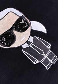 Un tessuto nero presenta un personaggio cartoon ricamato con un viso beige, capelli bianchi e occhiali da sole in paillettes nere; include contorni bianchi e un completo.