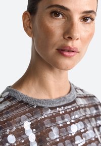 Gennemsigtig top med pailletter i en grå halsudskæring, der har cirkulære pailletter på en brun mesh-baggrund, hvilket fremhæver et tekstureret design.