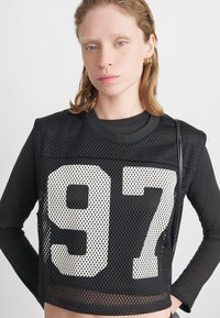 Schwarzes Mesh-Crop-Top mit großen weißen "97"-Grafiken, das über einem langärmeligen schwarzen Shirt liegt, mit nahtlosem Finish und leichtem Material.