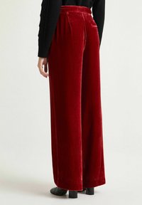Pantaloni ampi in velluto rosso con vita alta, pieghe sul retro e texture liscia. Scarpe nere visibili all'orlo.