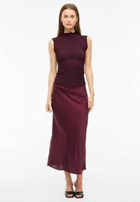 Burgundy ärmlös topp med texturerad tyg och en volangkrage, matchad med en slät, lång burgundy kjol. Platta svarta skor.