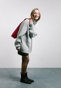 Suéter gris de canalé oversized, falda verde oscuro, botas negras hasta el tobillo y una mochila de tela roja. Texturas suaves y ajuste relajado.