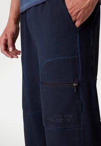 Marineblauwe joggers van zacht materiaal, met een ritszak en textuurelementen. Inclusief geprinte merkdetails aan de zijkant.
