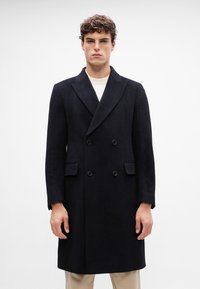 Cappotto nero doppiopetto con revers a punta, forma strutturata, tessuto testurizzato e due tasche anteriori, indossato sopra un maglione di colore chiaro.