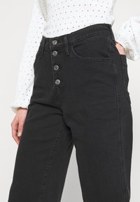 Vaqueros negros de talle alto hechos de mezcla de algodón con corte recto, que cuentan con cinco botones plateados y dos bolsillos delanteros. La textura es suave.