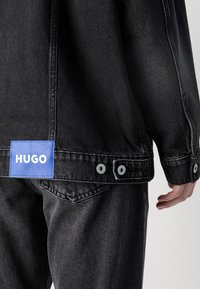Veste en jean noire et jean avec une étiquette bleue portant l’inscription "HUGO" sur le bas du dos, avec détails de boutons métalliques et une main sur le côté.
