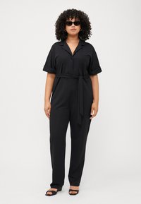 A woman with curly hair wears a black belted jumpsuit and dark sunglasses, standing against a plain white background.