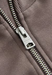 Tessuto grigio con una texture a coste, caratterizzato da una cerniera argentata. Il cursore della cerniera è inciso con "ALLSAINTS", evidenziando il dettaglio hardware.