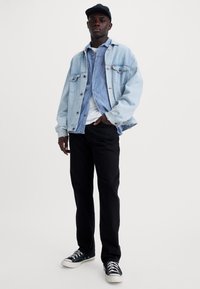 Lichtblauwe spijkerjack over een lichtblauw shirt, witte T-shirt, zwarte broek en zwarte sneakers. De pet is zwart, wat een casual accent toevoegt.