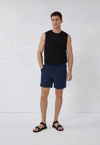 Čierny bezramienkový top, námornícke šortky s elastickým pásom a čierne sandále. Hladká tkanina, štandardný strih. Model stojí proti bielej stene.