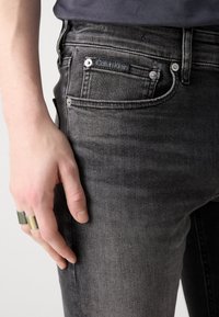 Černé džíny z denimu s úzkým střihem, na zadní kapse je štítek Calvin Klein a látka má jemné prošoupání.