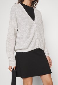 Světlomodrý pletený cardigan s výstřihem do V, volného střihu, se pěti knoflíky, kombinovaný s černými šaty. Měkká textura, jemný vzor.