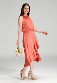 Giovane donna in un vestito satinado senza maniche color corallo e tacchi dorati, che tiene un piccolo bouquet, in piedi contro uno sfondo semplice e chiaro.